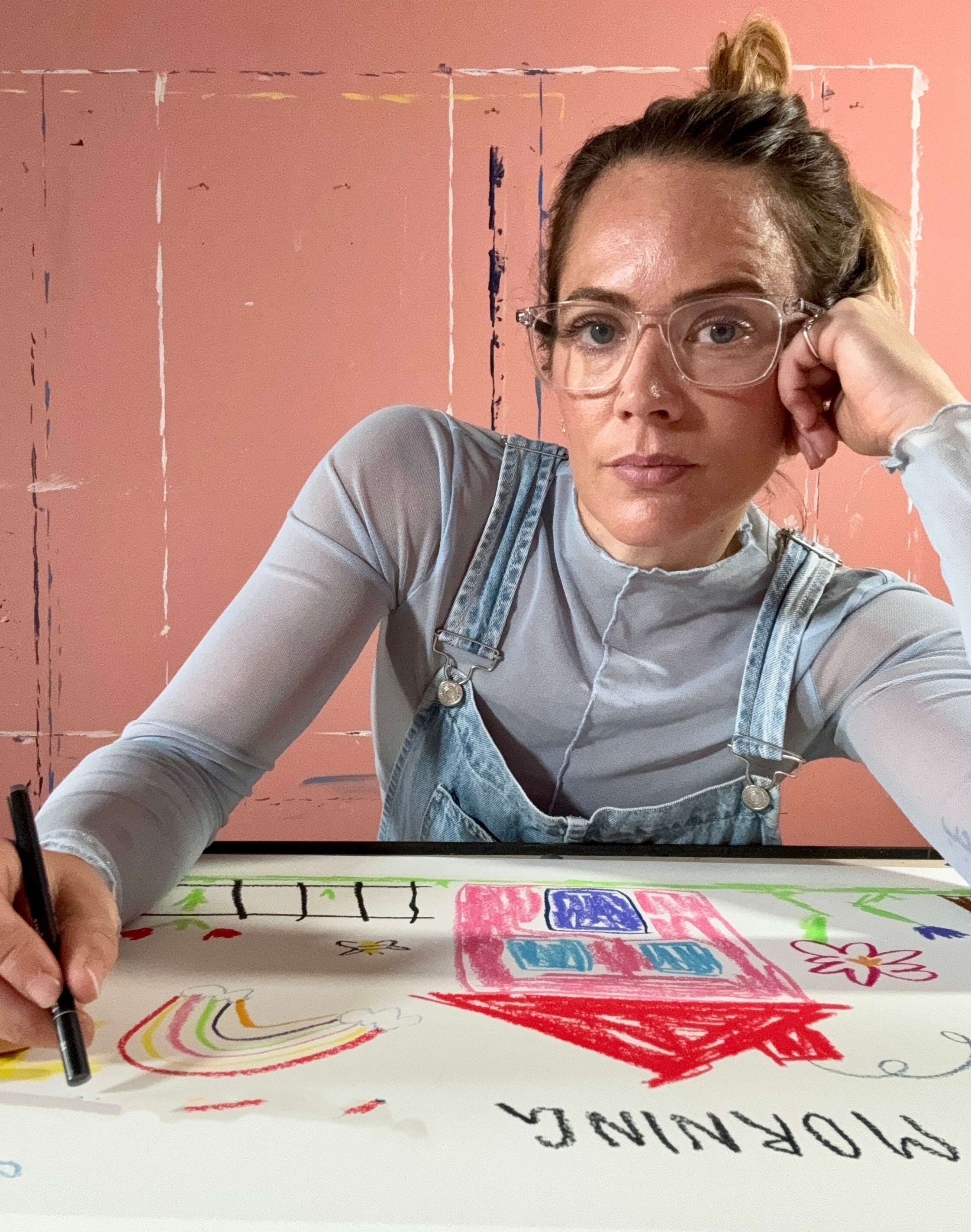 Artist with glasses and overalls sitting at a table creating a children's drawing on a pink wall background