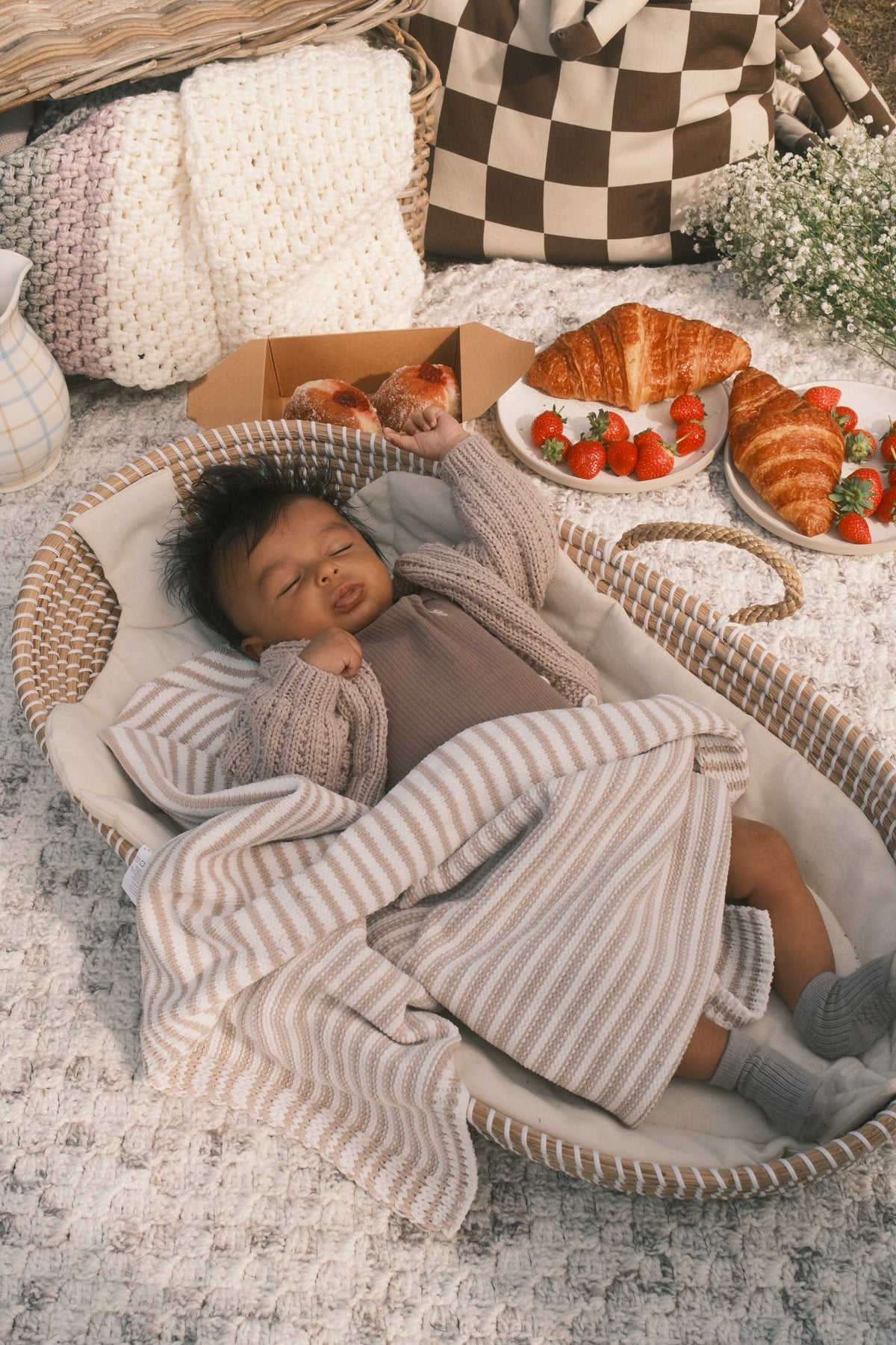 A baby wearing a brown knit outfit and a striped beige blanket is lying in a woven baby changing basket on a patterned rug, surrounded by breakfast items like croissants, strawberries, and a checkered bag in the background.
